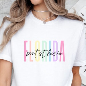 May include: White t-shirt with a rainbow-colored graphic that says "Florida port st. lucie" in black text.