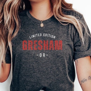 May include: Dark gray t-shirt with the text "LIMITED EDITION GRESHAM OR" in red. The shirt is a crew neck style and appears to be made of a soft material. The model is wearing light blue jeans and white sneakers.