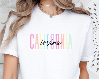 May include: White t-shirt with the words "CALIFORNIA irvine" printed in pastel colors. The text is centered on the front of the shirt. The shirt is made of a soft material.