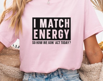 May include: A light pink t-shirt with the text "I MATCH ENERGY" in a black rectangle, followed by "SO HOW WE GON' ACT TODAY?" in a smaller font. The shirt is paired with denim shorts.