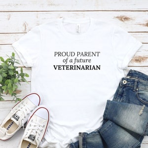 May include: White t-shirt with black text that reads "Proud Parent of a future Veterinarian". The shirt is laid out on a white wood background with a pair of white sneakers and blue jeans.