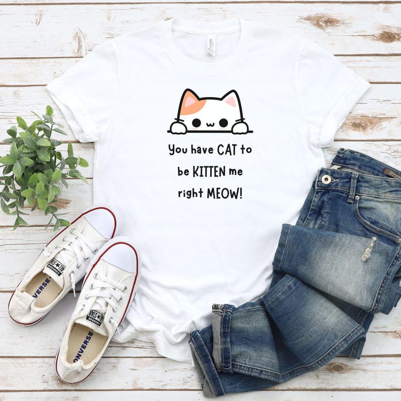May include: White t-shirt with a cartoon cat graphic and the text "You have CAT to be KITTEN me right MEOW!". The shirt is paired with blue jeans and white sneakers with red trim, creating a casual, everyday look.