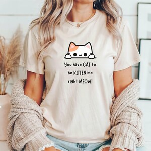 May include: Beige t-shirt with a cat graphic and the text "You have CAT to be KITTEN me right MEOW!". The cat graphic is black and white with orange accents. The shirt is short-sleeved.