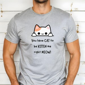 May include: A light gray t-shirt featuring a cartoon cat design with the text "You have CAT to be KITTEN me right MEOW!". The cat has white fur with orange accents. The shirt is made of a soft material.