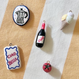 May include: Five small charms featuring a white oval with a black moka pot, a rectangular white tag with blue trim and the text CIAO BELLA, a black wine bottle with the text VINO, a red tomato, and a multi-coloured ice cream cone with a metal loop.