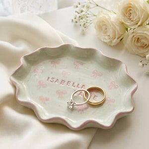 Op de afbeelding: Een ovale keramieken schaaltje met een geschulpte rand in lichtgroen, voorzien van een roze strikjespatroon en de naam ISABELLA in reliëf in het midden. Het schaaltje bevat een zilverkleurige ring met een steentje en een gouden ring.