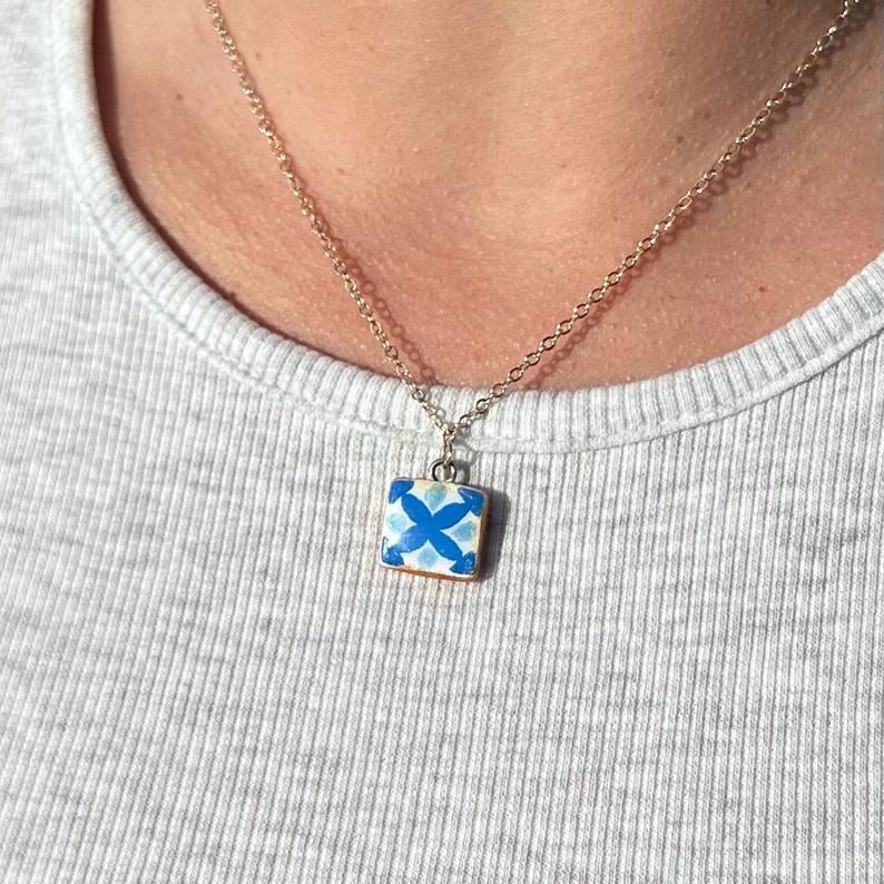 May include: A gold-toned metal chain necklace featuring a small, square pendant. The pendant displays a white and blue floral-inspired tile pattern with a symmetrical four-petal design centered on the surface, suspended from a simple metal bail.