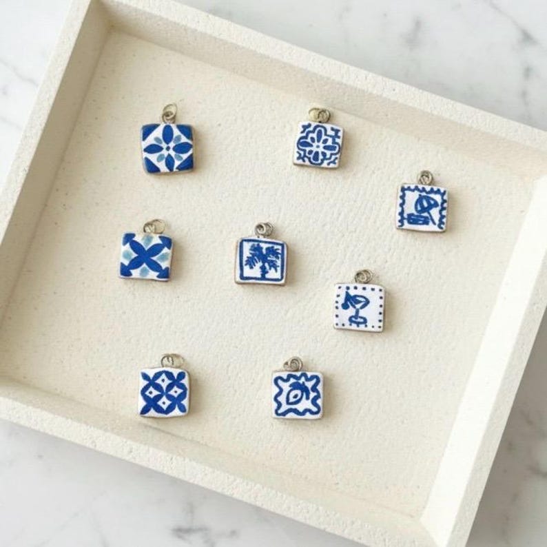 May include: Eight square charms featuring blue and white patterns, including geometric shapes, a palm tree, and a cocktail glass. Each charm has a small silver metal loop at the top for attachment, arranged on a light-colored square tray.