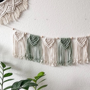 Puede incluir: Un tapiz de pared de macramé con una guirnalda de diseños en forma de diamante crema y verde alternados. La guirnalda está ensartada con pequeñas cuentas de madera y cuelga de una pared blanca. Una segunda pieza de macramé es visible en la esquina superior izquierda.
