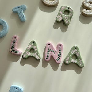 Puede incluir: El nombre "LANA" está escrito en letras de cerámica verde y rosa con diseños florales blancos y verdes.