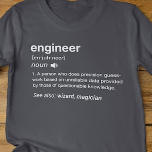 May include: A heather dark grey t-shirt with a white graphic print that defines the word "engineer" as a person who does precision guesswork based on unreliable data provided by those of questionable knowledge. The text also says "See also: wizard, magician".