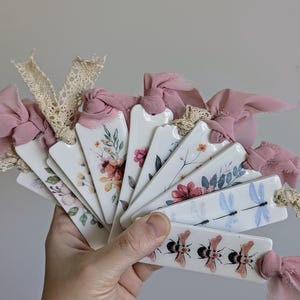 May include: A set of ceramic bookmarks featuring floral and insect designs. The bookmarks are white with watercolour illustrations of flowers, dragonflies, and bees. Each bookmark is tied with a pink ribbon.