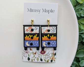 May include: A pair of black and gold dresser drawer earrings with floral, sun, and moon designs. The earrings are on a white card with the text "Mimsy Maple".
