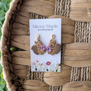 May include: A pair of brown squirrel earrings with purple flowers and gold accents. The earrings are on a white card with the text "Mimsy Maple you look gorgeous!"