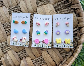 Puede incluir: Tres tarjetas con el texto "Mimsy Maple" en ellas. Cada tarjeta presenta un conjunto de tres pares de pendientes de arcilla de colores. Los pendientes tienen forma de zanahorias, pájaros, conejos, huellas de patas, setas, flores, huevos y mariposas.
