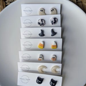 Puede incluir: Una colección de pendientes de botón con temática de Halloween, presentados en tarjetas blancas. Los diseños incluyen fantasmas, calaveras, sombreros de bruja, caramelos de maíz y gatos negros. Cada tarjeta lleva el texto "Mimsy Maple".