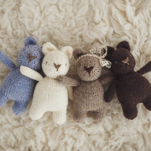 Puede incluir: Cuatro animales de peluche tejidos en diferentes colores: azul, blanco, marrón y marrón oscuro. Los animales están dispuestos en fila sobre una superficie blanca y esponjosa.