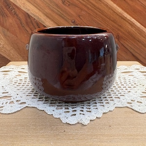 May include: A round, dark brown ceramic planter with a glossy finish and small side handles. The pot features a smooth, curved body and a wide opening at the top, showcasing a deep, reflective glaze that catches light across its rounded surface.
