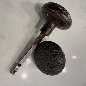 May include: A pair of dark brown, circular metal door knobs featuring an intricate, embossed floral pattern. One knob is attached to a long, square-shaped metal spindle, while the other knob is detached and positioned next to the spindle.