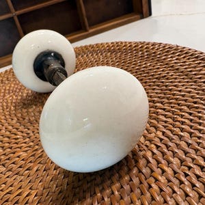 May include: A pair of round, white porcelain door knobs connected by a central metal spindle. The knobs have a smooth, glossy finish and a spherical shape, featuring a dark metal base where the handle meets the connecting rod mechanism.