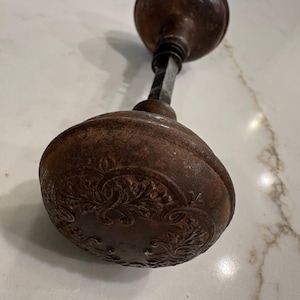 May include: A vintage round metal door knob featuring an intricate embossed floral pattern across its surface. The object has a dark, weathered brown patina and is connected to a central square metal spindle that joins two matching knobs together.
