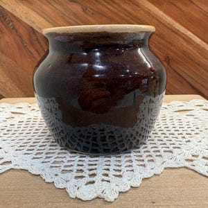 May include: A small, round ceramic pot with a glossy, dark brown glaze and a smooth, light tan rim. The vessel features a wide, bulbous body that tapers toward a narrow opening at the top, showcasing a simple, functional design with a reflective finish.