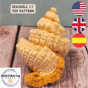 May include: A crocheted seashell in shades of yellow and gold. The seashell is made with textured stitches and features a spiral design. The text "Seashell 1.1 PDF Pattern" is visible in the upper left corner. The logo for Salt & Berry Knots is in the lower left corner.