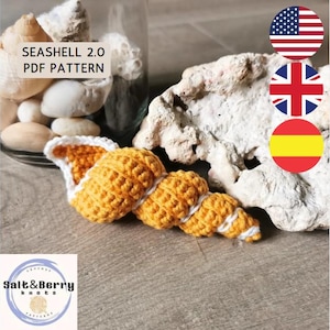 Puede incluir: Una concha de mar de crochet en amarillo y blanco, con el texto "Seashell 2.0 PDF Pattern" encima. La concha está sentada sobre una superficie de madera con un frasco de vidrio de conchas marinas en el fondo. La imagen también incluye las banderas de los Estados Unidos, el Reino Unido y España.