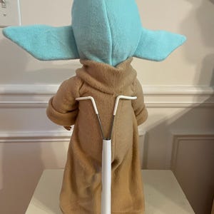 Peut inclure: Poup&eacute;e en peluche d'un personnage avec de grandes oreilles et une robe beige. La poup&eacute;e est sur un support blanc. La t&ecirc;te et les oreilles du personnage sont bleu clair. La poup&eacute;e est dos &agrave; la cam&eacute;ra.