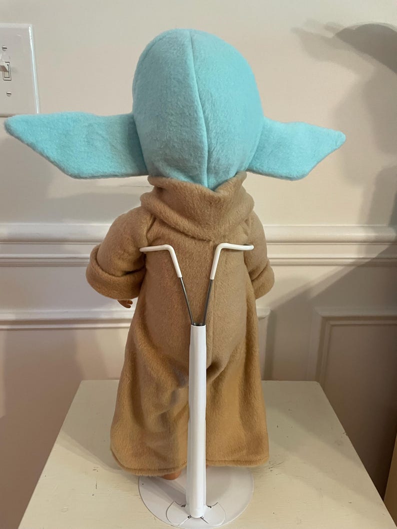Peut inclure: Poup&eacute;e en peluche d'un personnage avec de grandes oreilles et une robe beige. La poup&eacute;e est sur un support blanc. La t&ecirc;te et les oreilles du personnage sont bleu clair. La poup&eacute;e est dos &agrave; la cam&eacute;ra.