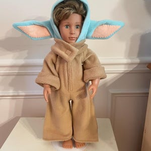 Peut inclure: Une poup&eacute;e v&ecirc;tue d'une combinaison beige et d'une capuche bleu clair avec de grandes oreilles. La capuche a des d&eacute;tails d'oreilles int&eacute;rieures roses. La poup&eacute;e est pos&eacute;e sur une surface blanche.