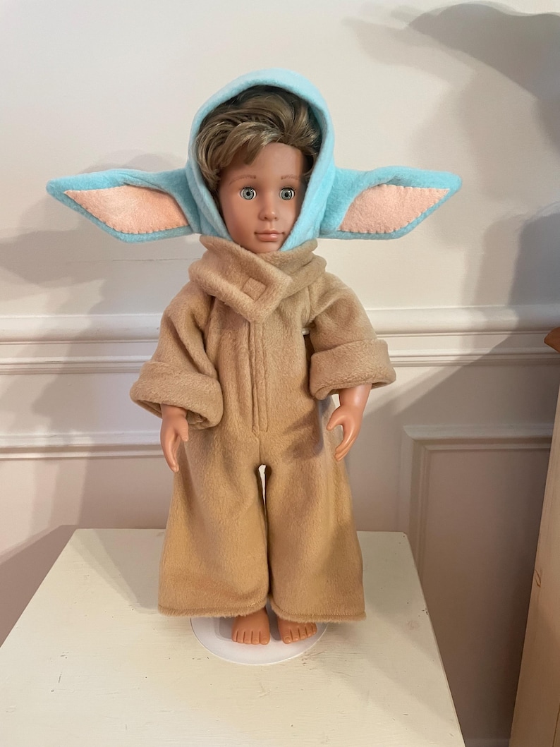 Peut inclure: Une poup&eacute;e v&ecirc;tue d'une combinaison beige et d'une capuche bleu clair avec de grandes oreilles. La capuche a des d&eacute;tails d'oreilles int&eacute;rieures roses. La poup&eacute;e est pos&eacute;e sur une surface blanche.