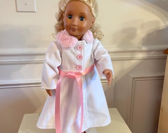 May include: A doll with blonde curly hair and blue eyes wearing a white coat with a pink fur collar and pink ribbon belt. The coat has pink buttons and the doll is wearing pink shoes. The doll is standing on a white surface.