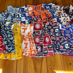 May include: A collection of colorful dresses featuring various college team logos and colors. The dresses are made of fabric with repeating patterns of team names, mascots, and logos. The dresses are in various colors, including red, blue, orange, and yellow.