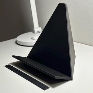 Könnte beinhalten: Ein schwarzer dreieckiger Telefon- oder Tablet-Ständer mit einer strukturierten Oberfläche und einem dünnen schwarzen Streifen, der an der Unterseite befestigt ist.