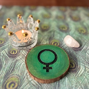 May include: A green wooden circle with a black female symbol painted on it. The circle is sitting on a table with a glass crown candle holder and a white stone.