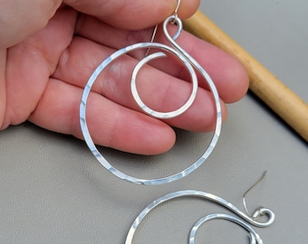 May include: A pair of silver hoop earrings with a hammered finish. Each earring features a smaller circle that is twisted and wrapped around the larger circle.