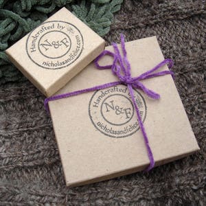 May include: Two small, brown cardboard boxes with a purple ribbon on one. Both boxes have a black and white logo with the text "Handcrafted by N&F nicholasandfelice.com".