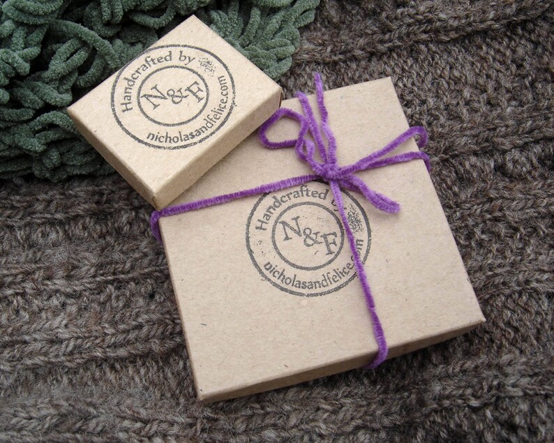 May include: Two small, brown cardboard boxes with a purple ribbon on one. Both boxes have a black and white logo with the text "Handcrafted by N&F nicholasandfelice.com".