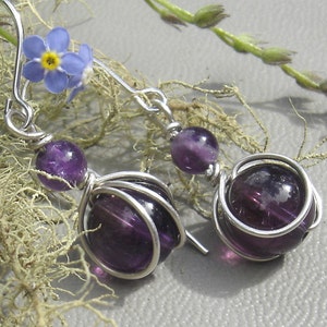 May include: A pair of silver earrings with purple amethyst stones. The stones are wrapped in silver wire and dangle from silver hooks.
