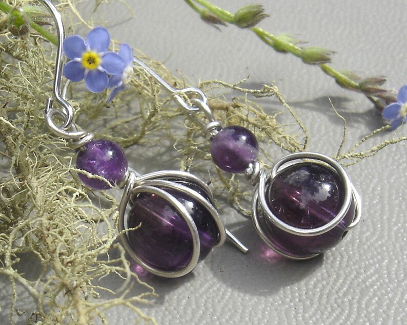 May include: A pair of silver earrings with purple amethyst stones. The stones are wrapped in silver wire and dangle from silver hooks.