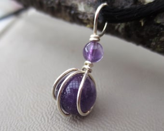 Könnte beinhalten: Ein handgefertigter Anhänger mit einem großen, runden, tiefvioletten Amethyststein, der in einem silbernen Drahtkäfig eingeschlossen ist. Eine kleinere Amethystperle sitzt darüber, verbunden durch Silberdraht. Der Anhänger hängt an einer schwarzen Kordel.