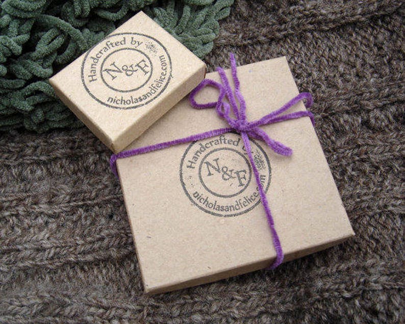 May include: Two small, brown cardboard boxes with purple ribbon. The boxes are stamped with the text "Handcrafted by N&F nicholasandfelice.com".