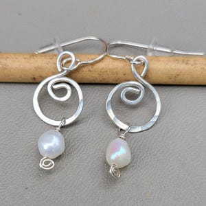 May include: A pair of silver-toned wire earrings featuring a circular spiral design with a hammered texture. Each earring is accented with a single white, iridescent freshwater pearl dangling from the bottom, secured with a small wire coil.
