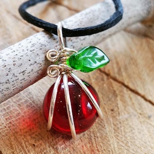 以下が含まれることがあります： 銀色のワイヤーで包まれた赤いガラスのリンゴのペンダントと、緑色のガラスの葉。ペンダントは黒いコードに付いています。