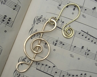 May include: A gold-colored metal treble clef bookmark. The bookmark is shaped like a treble clef and is lying on a sheet of music.