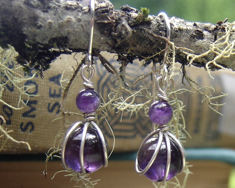 May include: A pair of silver earrings with amethyst gemstones. The amethyst is wrapped in silver wire and hangs from a silver hook.