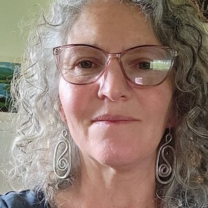 May include: A woman with gray curly hair wearing glasses and silver earrings. The earrings are shaped like a figure eight.