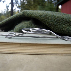 May include: A silver metal brooch with a spiral design. The brooch is lying on a book with a green fabric background.