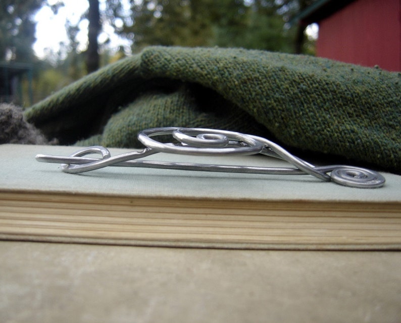 May include: A silver metal brooch with a spiral design. The brooch is lying on a book with a green fabric background.